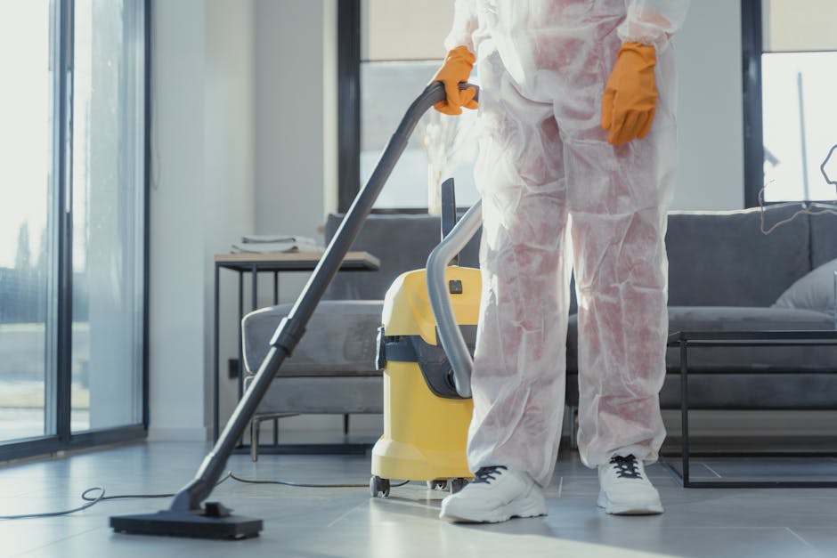 A professional cleaner dressed in full protective gear, including a white coverall, face mask, goggles, and blue gloves, is performing surface cleaning and sanitisation on a white, glossy round dining