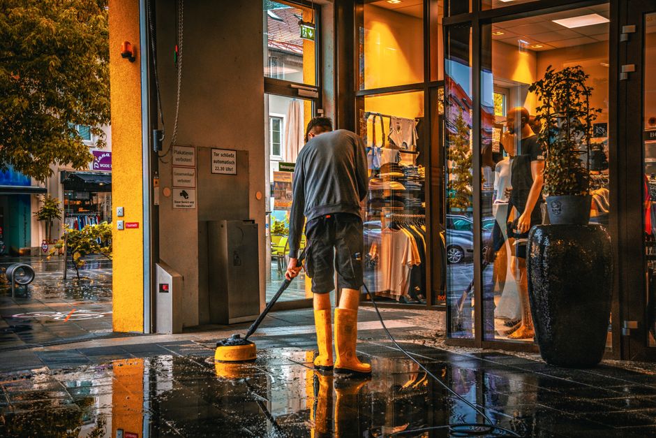 A professional window cleaner working outside a large glass storefront window, cleaning the surface with a squeegee and a cloth. The glass panels are partially wet, with visible water droplets and str